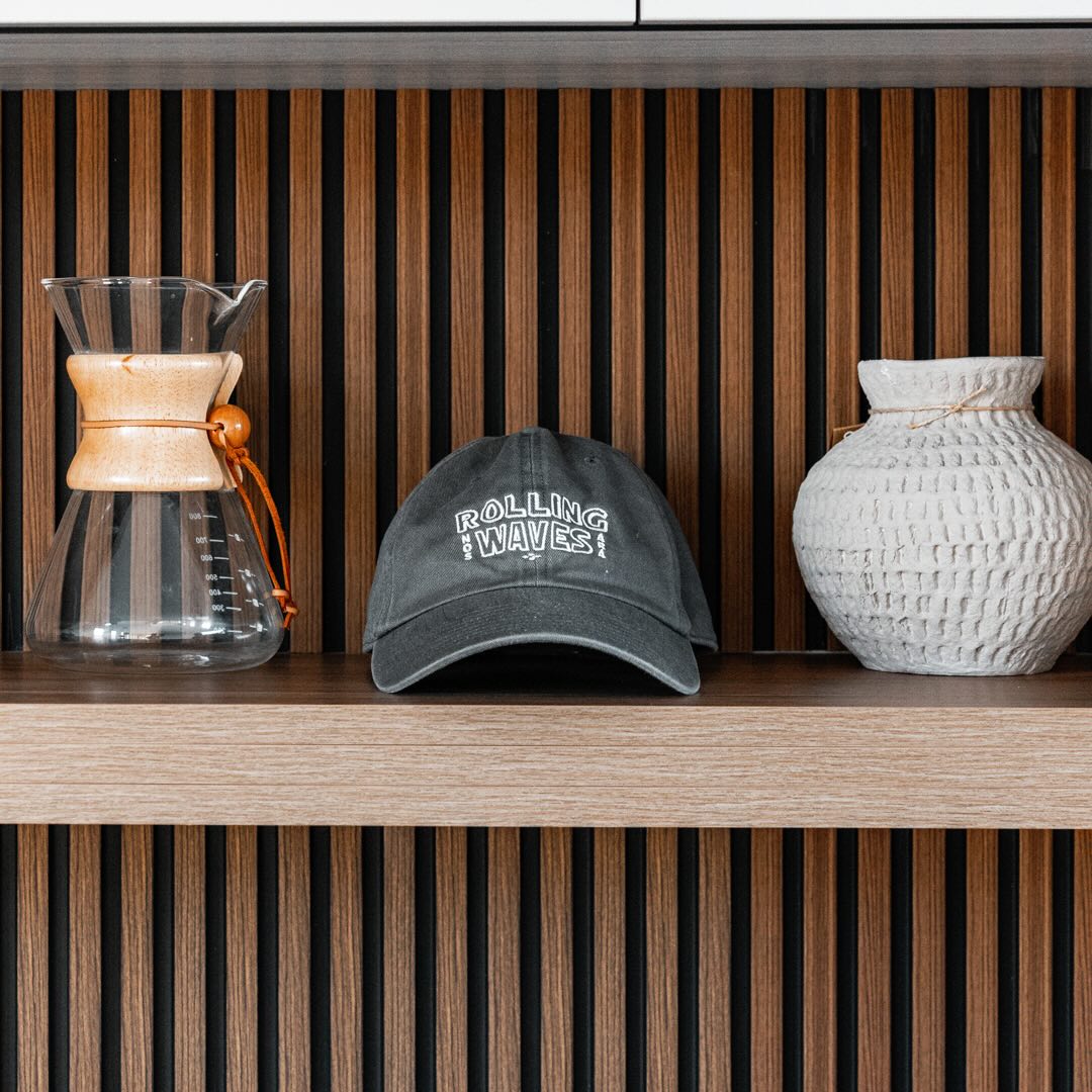 Cap with 'Rolling Waves' text on a shelf with a Chemex coffee maker and textured vase against a striped background