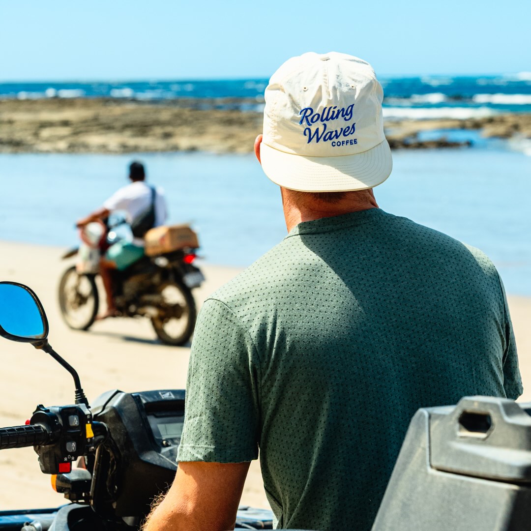 Man wearing a 'Rolling Waves Coffee' cap sitting on a quad on the beach.